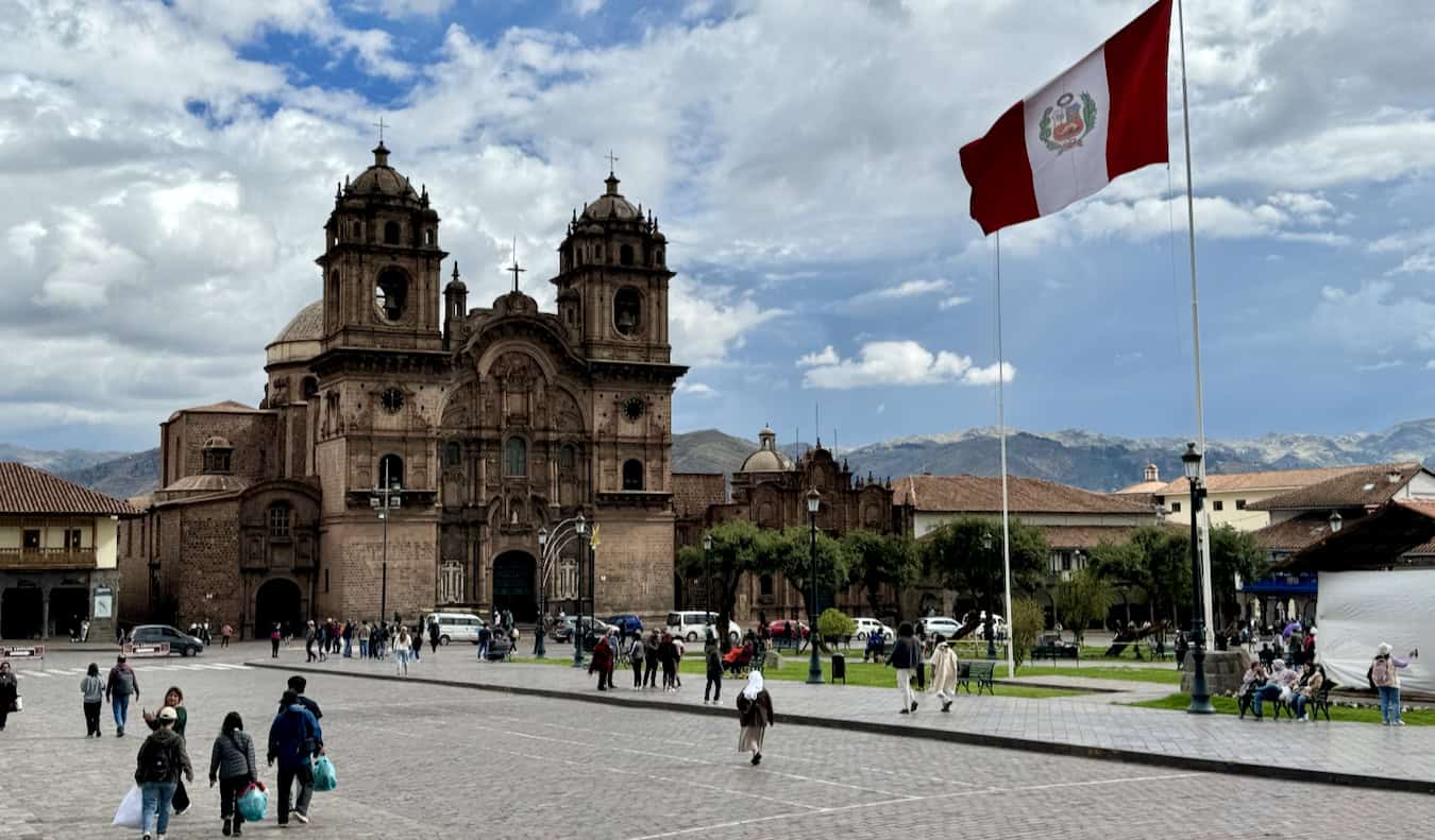 A sunny day in Peru with a huge flag waving in the breeze with historic buildings nearby